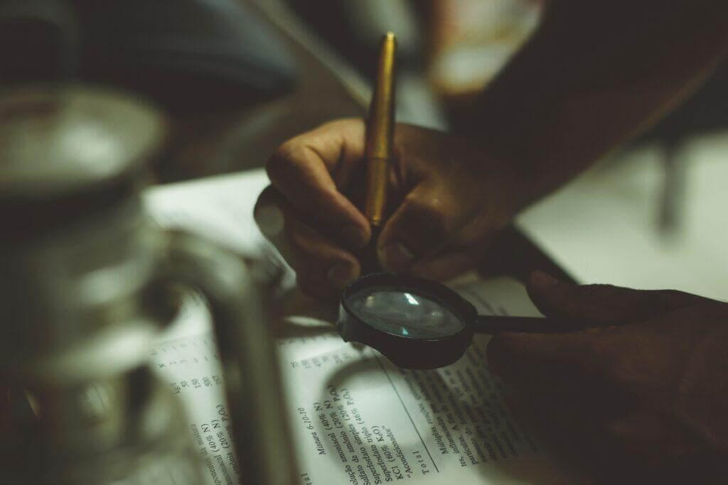 Person reviewing a book with magnifying glass and pen.
