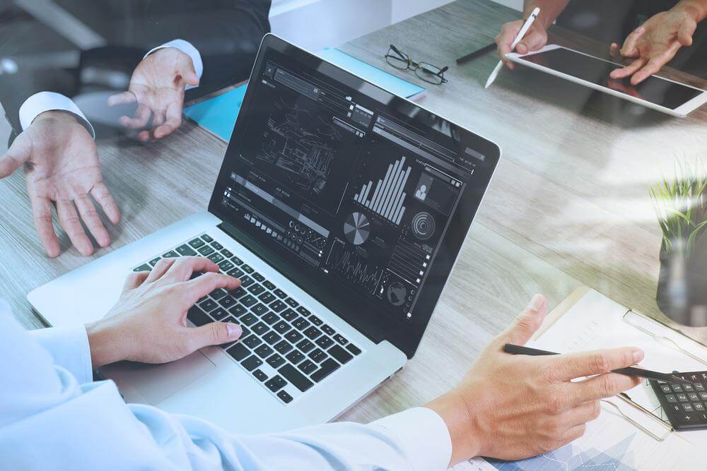 Business investment firm documents on office table with smart phone and laptop and graph financial with social network diagram and three colleagues discussing data in the background.
